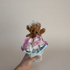 eRamisHere brown teddy bear wearing a tiara holding a bouquet of pink and white roses with pearls.
