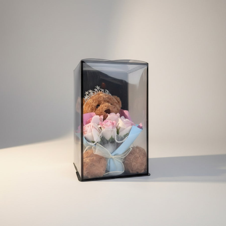 eRamisHere brown teddy bear with tiara holding a bouquet of pink and white roses in a clear gift box.