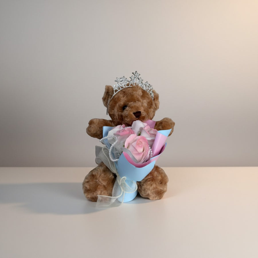 Brown teddy bear wearing a silver tiara, holding a bouquet of pink and white roses with pearl accents.